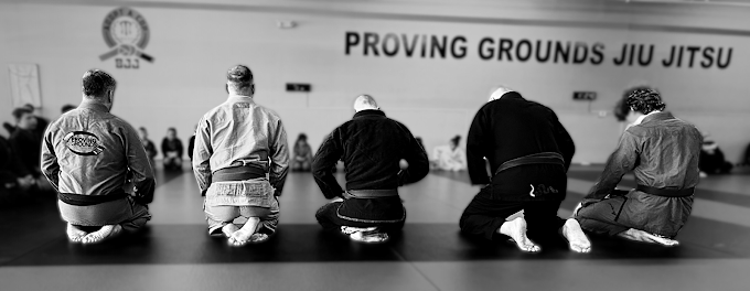 Black-and-white photo of BJJ students kneeling in a line on the mats facing the Proving Grounds Jiu Jitsu wall.