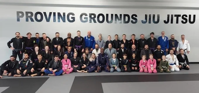 Large group of adult and kids BJJ students in gis posing in front of the Proving Grounds Jiu Jitsu wall.